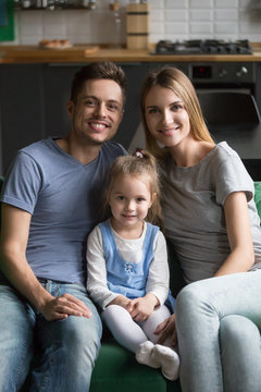 Vertical portrait of happy smiling family with preschool daughter, mother and father sitting on sofa at home together with little girl, kid sitting between parents, look at camera