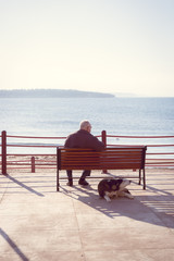 A man sitting on the bench alone with a dog lying down on his behind