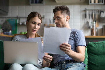 Upset man and woman sitting together on sofa, using laptop, wife and husband checking documents,...