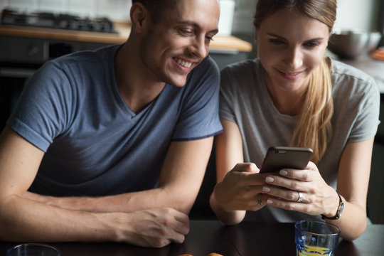 Young smiling couple looking at smartphone screen, watching funny video in social network, good photos, man and woman having breakfast together at home, checking email, online shopping, free weekend