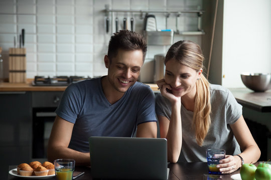 Happy young couple looking at laptop screen in kitchen together, laughing, having fun at home, using computer for reading online funny news, watching video, chatting in social networks