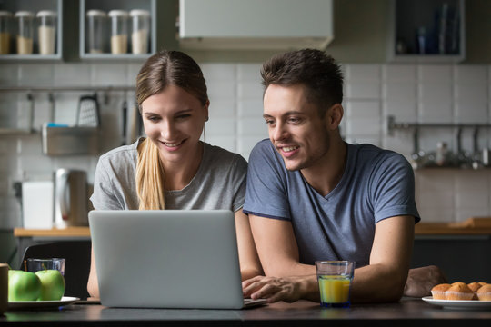 Happy young smiling couple looking at laptop screen in kitchen together, spending time at home together, using computer for reading online good news, watching video, chatting in social networks