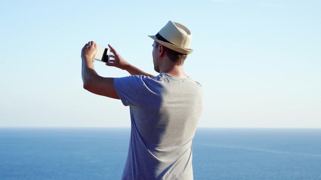 Young handsome man relaxing at edge of cliff take pictures on cellphone. Male in hat enjoying breathtaking view of blue Mediterranean sea making photos on mobile phone