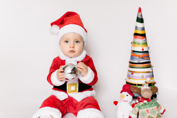 Little girl wearing santa claus red dress sitting on the floor holding small Christmas ball and smiles over white background,Christmas concept.