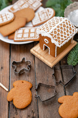 Cute little house. A lot of ginger biscuits in different form on brown wooden table. Decorated with white sweet glaze. Christmas mood, winter morning. Fir branches