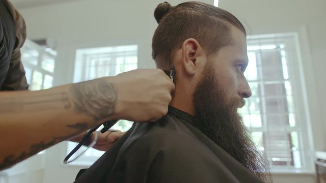 Bearded man getting trendy haircut at salon. Hairstylist giving stylish haircut to client at barber shop.