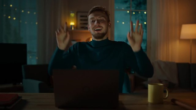 In the Evening: Happy Young Man Dances while Sitting at His Desk, He Uses Laptop and Listens to a Music.