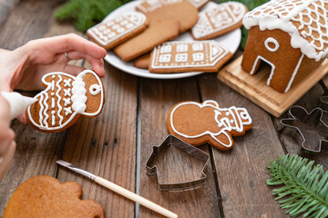 Icing of Christmas bakery. Woman decorating honey gingerbread cookies on wooden brown table. closeup, copy space. Blank biscuit gingerbread house, ready to decorate.