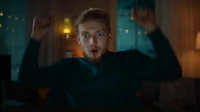 In the Evening: Man Sitting at His Desk and Using Laptop Gets Surprised, then Happy and Celebrates Win with Dance. In the Background Cozy Living Room.