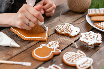 A young girl decorates ginger cookies Christmas winter morning. Woman draws Icing on honey gingerbread house. Wooden brown table. copy space. Blank biscuit gingerbread house, ready to decorate.