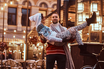 Young romantic couple enjoying spending time together at Christmas on street decorated with beautiful lights. Happy young guy holding his girls in his arms.