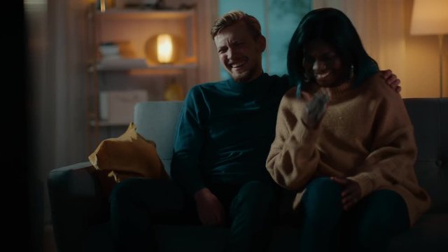 Happy Diverse Young Couple Watching Comedy on TV while Sitting on a Couch, they Laugh and Enjoy Show. Handsome Caucasian Boy and Black Girl in Love Spending Time Together. Moving Camera Shot.