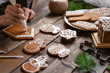 Icing of Christmas bakery. Woman decorating honey gingerbread cookies on wooden brown table. closeup, copy space. Blank biscuit gingerbread house, ready to decorate.
