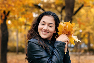 Pretty woman is posing with bunch of maple's leaves in autumn park. She's closing eyes and dreaming. Beautiful landscape at fall season.