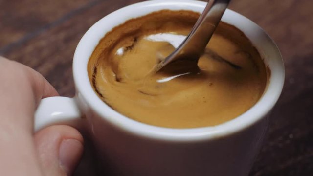 Man hand holding white coffee cup with freshly brewed espresso with foam, pouring sugar down and mix up with a spoon. Person making and drinking freshly made black coffee in the morning. Close-up.