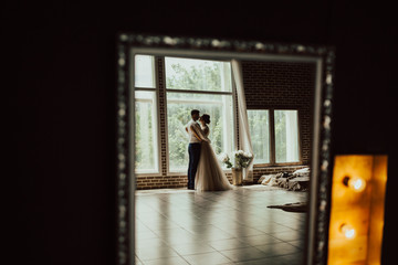 Young man gently embracing his bride and looking with love into her eyes.Newlyweds at wedding day.Happy luxury bride and groom standing at window light in rich room, tender moment.