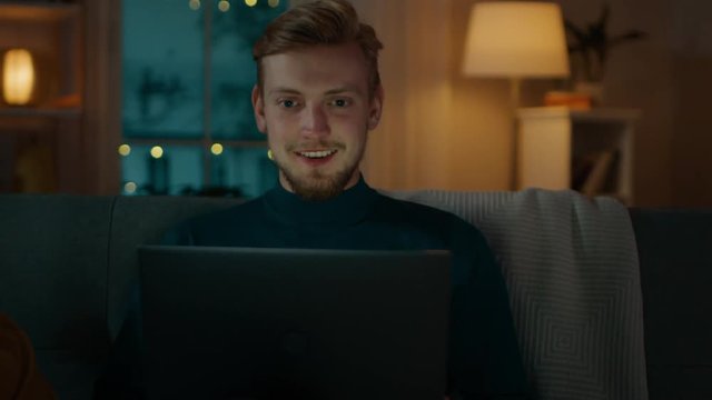 Portrait of a Handsome Young Man Sitting on a Couch Using Laptop to Make a Video Call, Says Hello, Waves His Hand. In the Evening Man Talks with Relatives and Friends Using Computer Webcam.