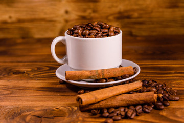 White cup filled with coffee beans and cinnamon sticks on wooden table