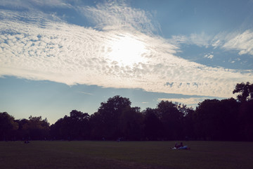 Sun behind white puffy clouds, shinning above the the trees in a public park, sunset time