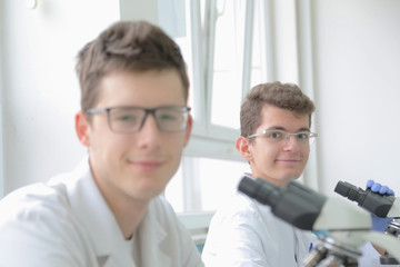 Fototapeta na wymiar Two Young male scientists looking through a microscope in a laboratory doing research, microbiological analysis, medicine. Smilling into camera.