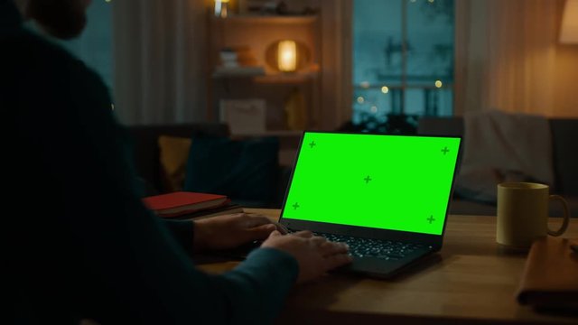 Man Sitting at His Desk Works on a Laptop with Green Chroma Key Screen. Late at Night in His Living Room Man Uses Notebook Computer. Side View Zooming on Screen.
