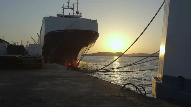 wide still shot, fine sunset evening, front view of a docked ship, tied by large hanging ropes, sky, ball of bright yellow sun above horizon hill ready to set, vivid reflections on grey calm water