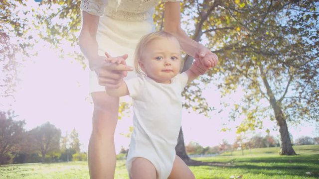 Young Caucasian mother supporting her little baby son as he practices his first steps on the grass 