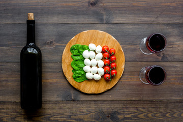 Italian cuisine, food concept. Italian flag made of mozzarella, tomatoes, basil on wooden cutting board near glass of red wine and wine bottle on dark wooden background top view