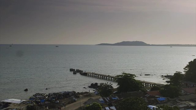 wide aerial, still dark shot, stony Conakry beach from land, light-grey water, slanting jetty, dull sky, stones.  boulders, silhouettes of fishing canoes, rusty roof-tops of slum houses, beach roa