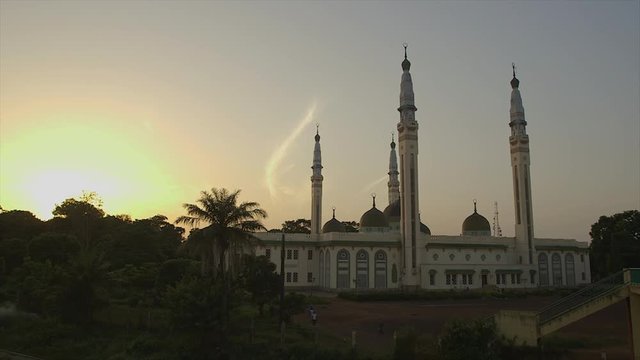 Medium still shot, decorated, cream painted Grand Mosque of Conakry, Guinea, tall, minarets, bold domes. Behind silhouettes of thick leafy coastal trees, palm leaves, sun setting, low darkness, a ring