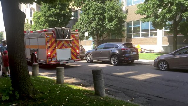 Steady panning shot of a fire brigade rushing to an accident spot with alarm lights blinking, on a road with cars parked on both sides. Man trying to cross the road with his sons, on an afternoon
