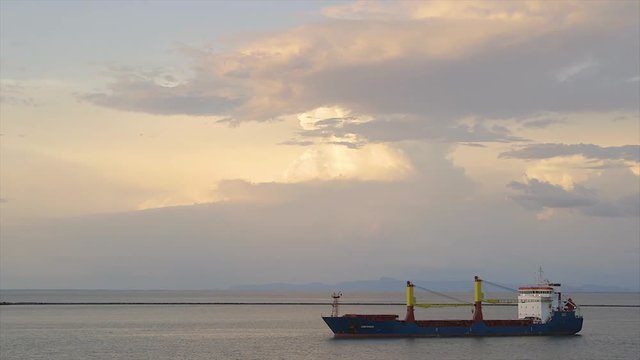 Wide still evening shot, vast, grey, calm, quiet Conakry port's Ocean water; thick dark-bright sunset sky clouds, large blue, brown empty cargo ship with yellow painted deck cranes stays stationary