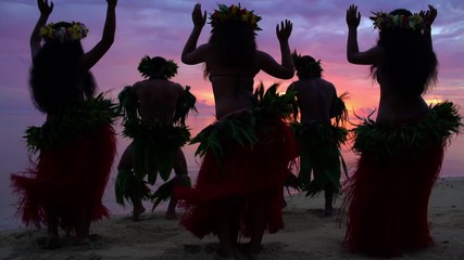 Barefoot Tahitian males in warrior dress on the beach at sunset with females in hula skirts and flower headdress performing a traditional dance French Polynesia South, Pacific,