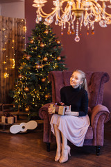 Obraz na płótnie Canvas Beautiful young woman dressed in a sweater and skirt sitting on the arm chair next to the christmas tree with gift box in hands.