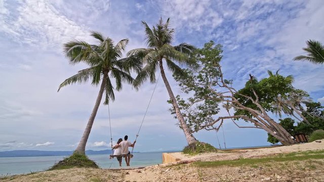 A young couple swinging on a swing on a tropical beach. Couple in love on a beach swing by the sea in southeast Asia.