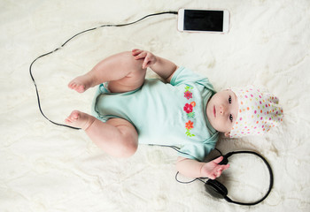 little beautiful newborn baby lying on the bed listening to music and next to the headset with the phone close-up