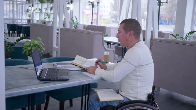 e-learning of disabled, ill student male on wheel chair uses modern computer technology to distance studying from online lecture and books making notes in notebook in cafe