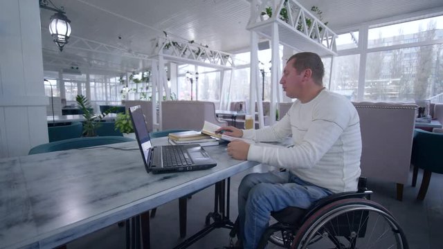 successful student crippled man on wheelchair using laptop computer for video education to learn from online teaching making notes in notebook in cafe