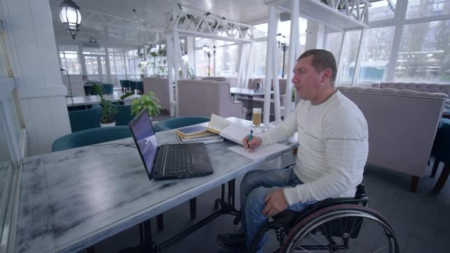 Successful invalid restaurant owner man on wheelchair uses modern computer technology for management and development business ideas indoors