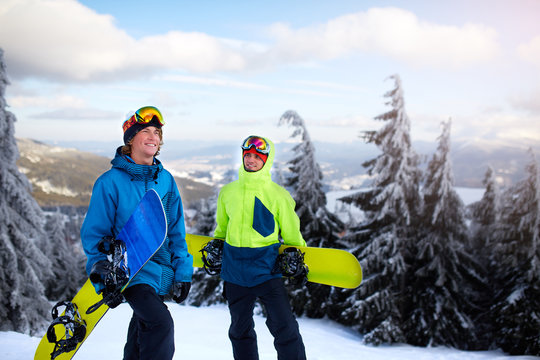 Two snowboarders walking at ski resort. Friends climbing to mountain top carrying their snowboards through forest for backcountry freeride and wearing reflective goggles, colorful fashion clothes.