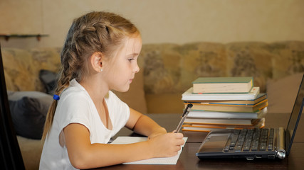 Side view of little girl with braids sitting at table with stack of books watching laptop and doing homework