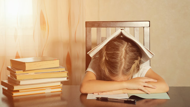 Little Girl With Textbooks Looking Tired And Leaning On Hands Sitting At Table And Studying