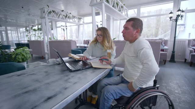 education courses for disabled, Smart aching student mature men in wheelchair with tutor female during home education using modern computer technology in restaurant