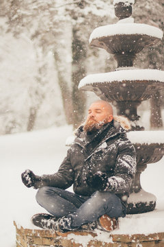 Bearded man meditates sitting on a snowy fountain while is snowing - peace of mind and healthy lifestyle concept
