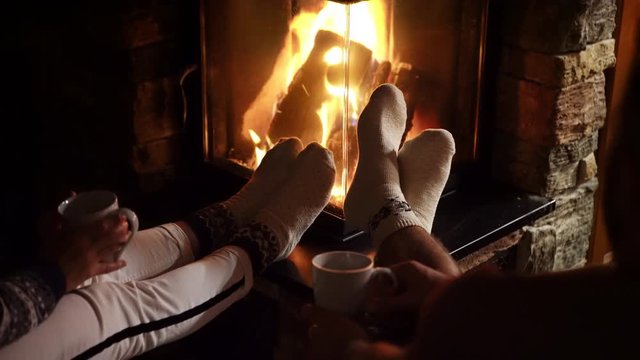 Young couple warming their feet pulled with Christmas ornamented socks near the fireplace fire and drinking the tea. Cozy Christmas Home concept 4K Footage