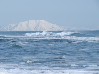Pacific Ocean, waves and views of the snow-covered hill in winter in sunny weather in Kamchatka, Russia