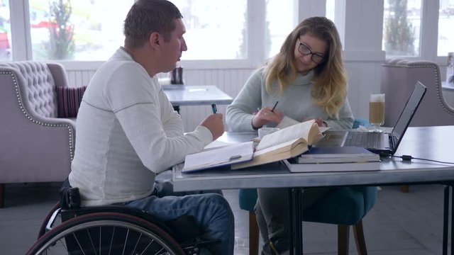 individual education for crippled, tutor women spectacles holds lesson for disabled men on wheelchair using a laptop computer and books in restaurant