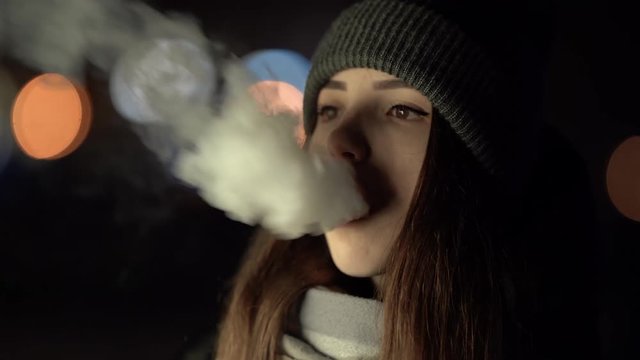 Close portrait of a girl smoking vape outside in the evening. Nice colorful bokeh in front of the evening city.