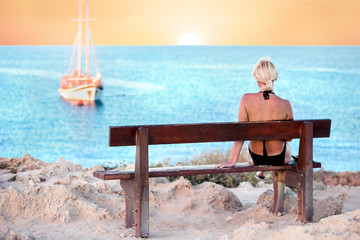  woman in swimsuit sitting on the bench looking sea, sunset, sailboat. relax travel concept.