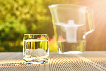 Clean glass of a clear water with a water filtration jug lit by sun rays in a green garden in warm summer day